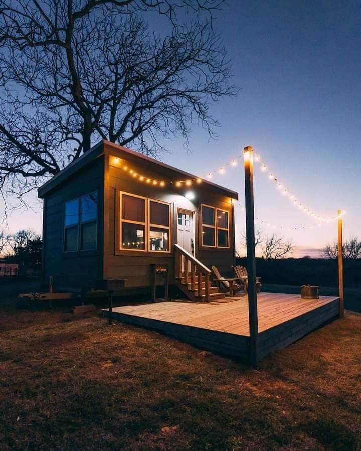 Cozy tiny house on a wooden deck with string lights at dusk, surrounded by bare trees.